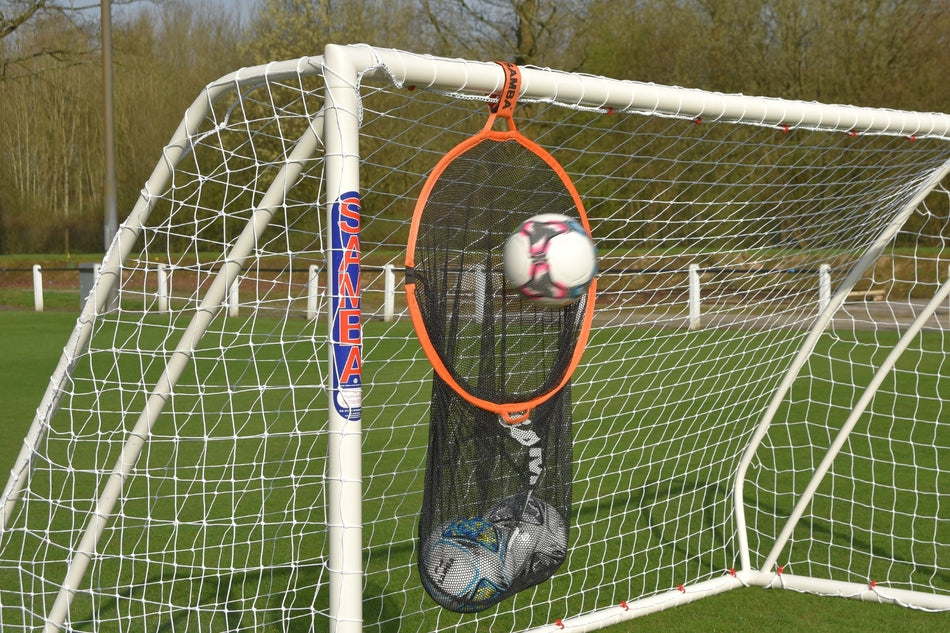 A Samba Sports orange training target is fixed to the goalpost, with extra balls stored in a Samba 2 in 1 Target Bag below—all on a grassy field.