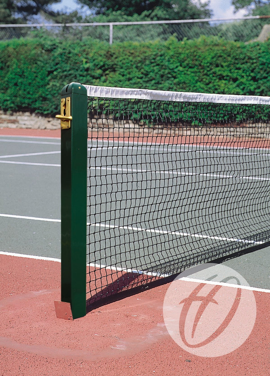 A close-up of Harrod Sport Aluminium 80MM Square Tennis Posts with net on an outdoor hard court, green fencing, and trees in the background beneath a partly cloudy sky.