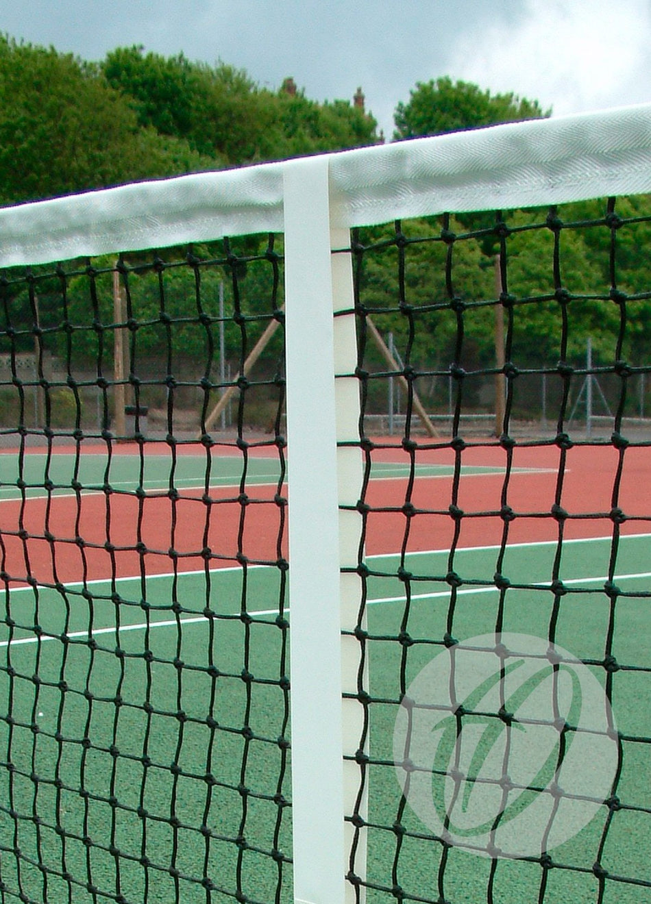 Close-up of a Harrod Sport Woven Polyester Centre Tape with brass eyelets on a tennis net, set on an outdoor court with green and red surfaces. Trees and a cloudy sky are visible in the background.
