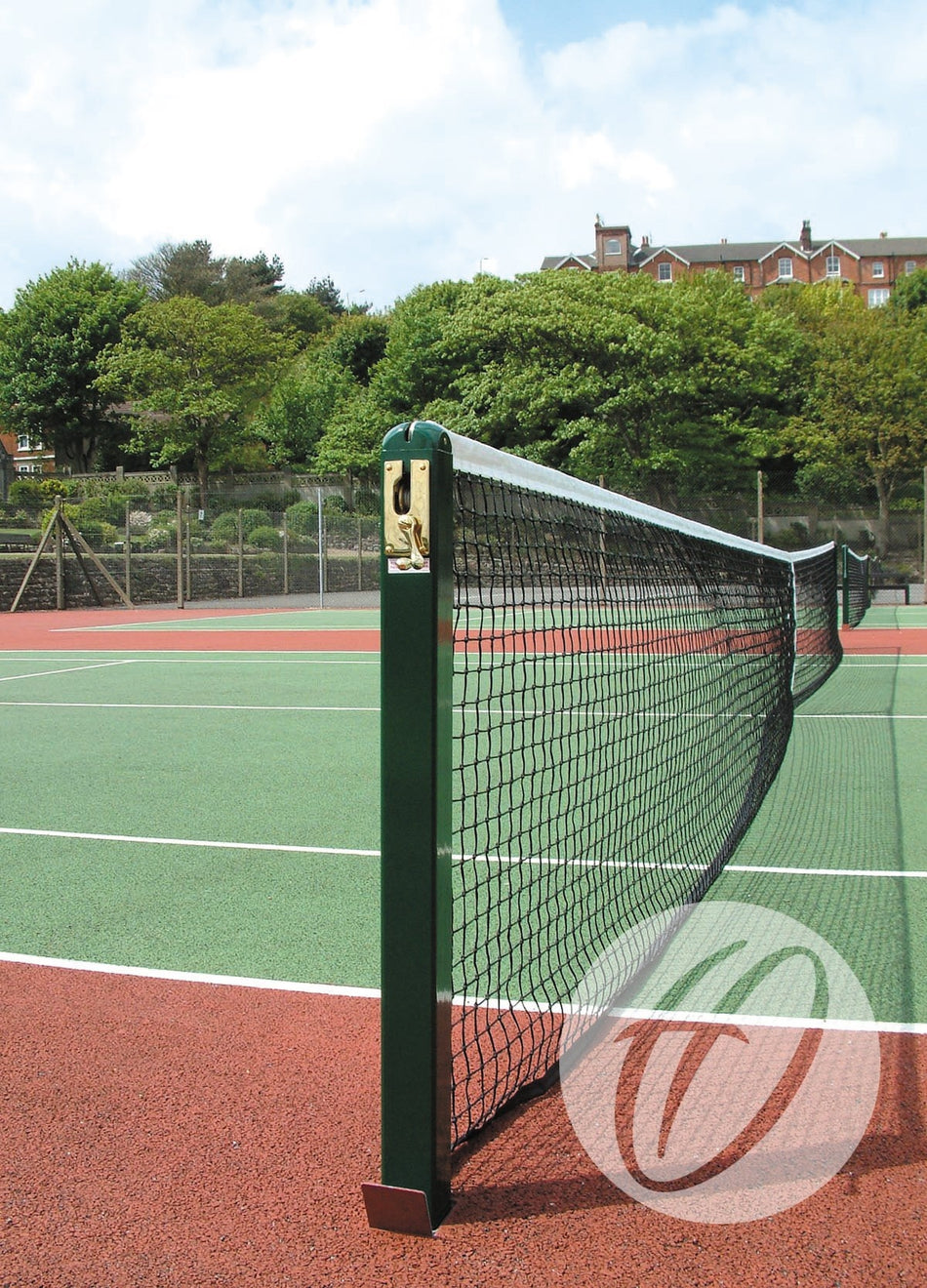 A close-up of the Harrod Sport Spare S8 Winder Tennis Post with a heavy-duty brass winder on an outdoor court featuring a green surface and red borders, surrounded by trees and buildings under a partly cloudy sky.