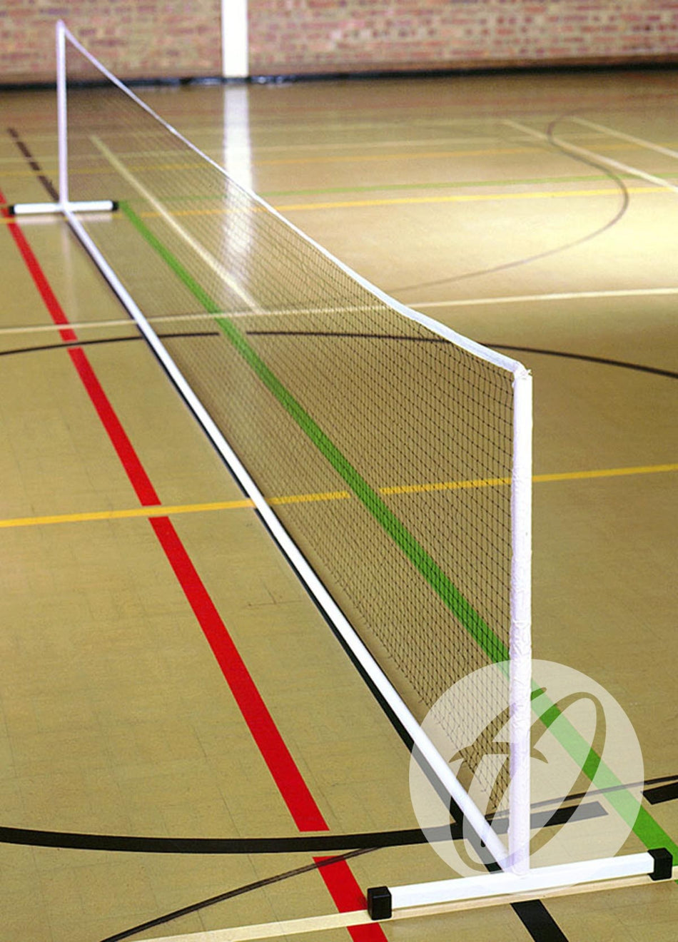 A Harrod Sport Freestanding Practice Mini Tennis Net is set up indoors on a gym floor with colorful court lines and a brick wall behind, supported by sturdy Harrod Sport Mini Tennis Posts.