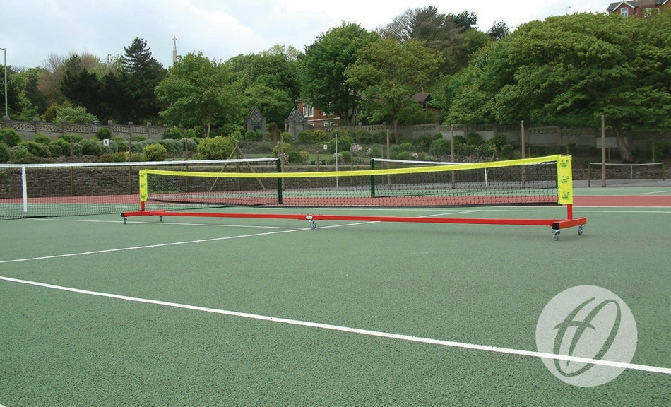 The Harrod Sport Wheelaway Mini Tennis Posts are set up on a green outdoor tennis court surrounded by trees and shrubbery, with buildings visible in the background.