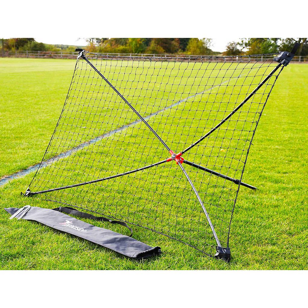 The Precision Training Quick Setup Portable Rebounder (5ft x 3ft) with an X-shaped frame and adjustable angle stands on a grassy field. Its black carrying bag is in front of the net, with trees and a fence in the background.