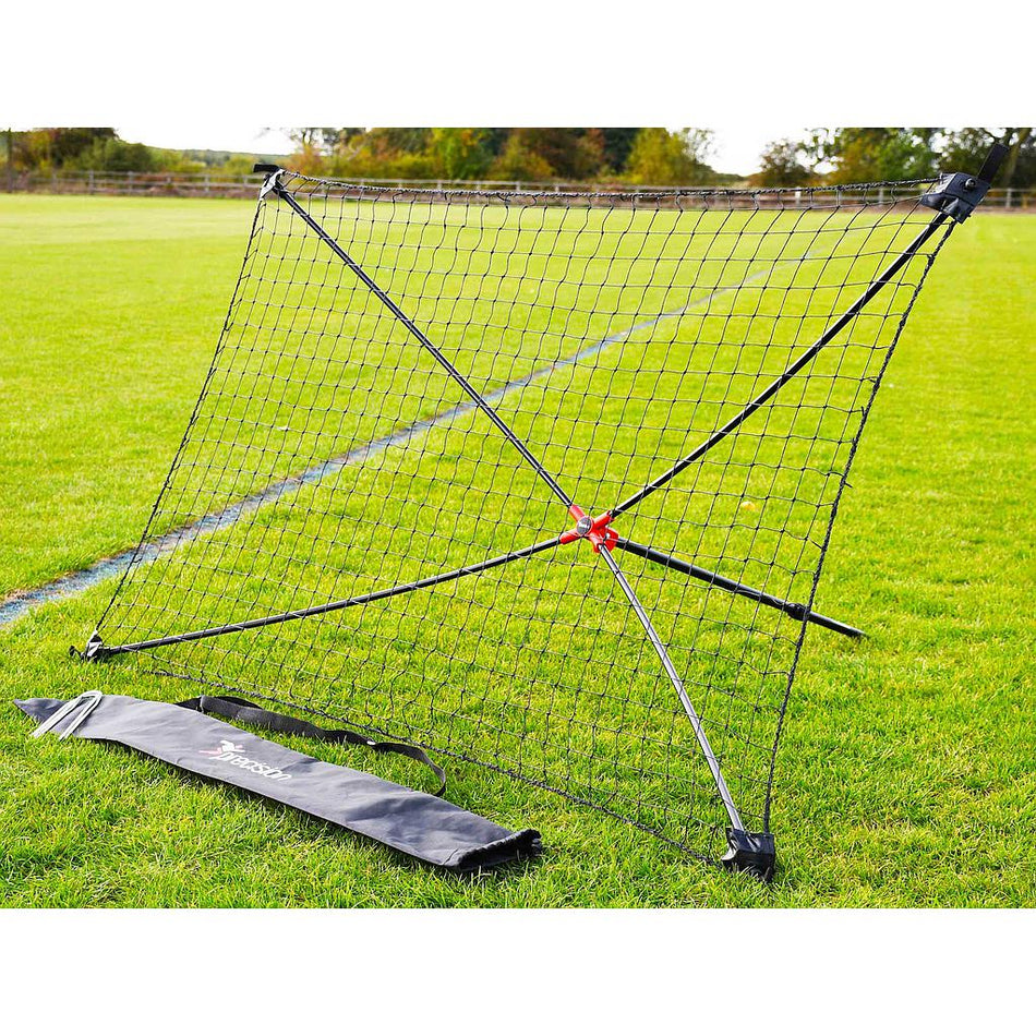 The Precision Training Quick Setup Portable Rebounder (5ft x 3ft) with an X-shaped frame and adjustable angle stands on a grassy field. Its black carrying bag is in front of the net, with trees and a fence in the background.