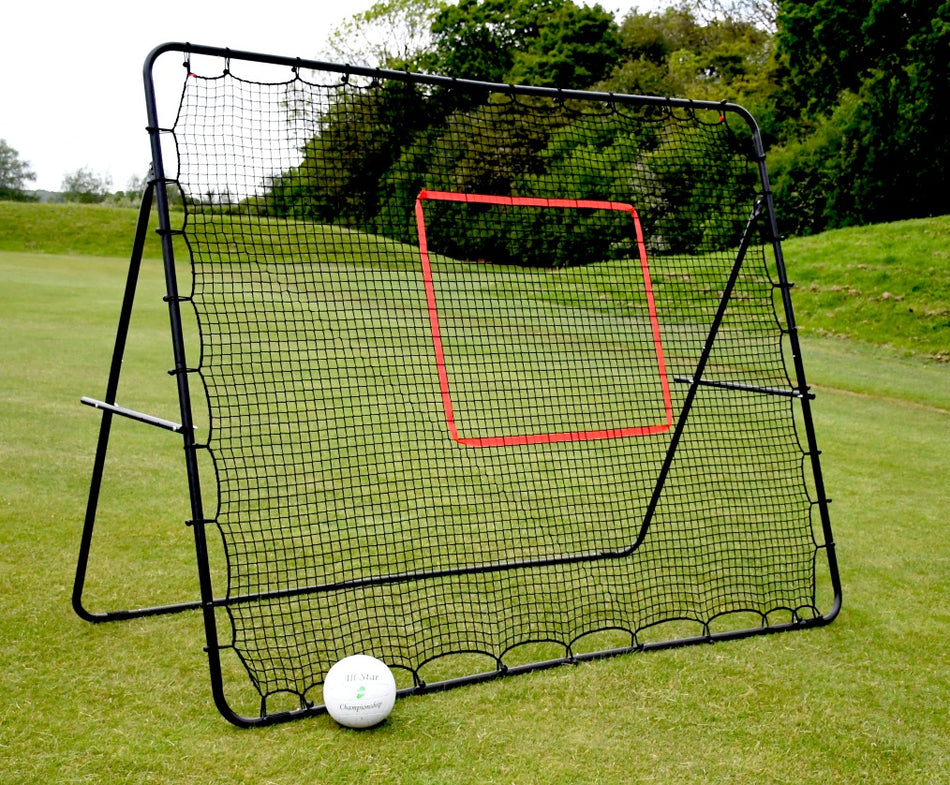 The Precision Training Precision Pro Jumbo Rebounder, measuring 9ft x 7ft with a red target area, stands on grass with a white soccer ball in front. Trees and bushes in the background show its multi-sport versatility.