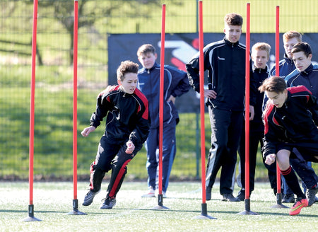 A group of boys in sportswear weave between Precision Training’s Precision Boundary Poles (Set of 12) with ground spikes on a grassy field, practicing lateral drills while others wait for their turn.
