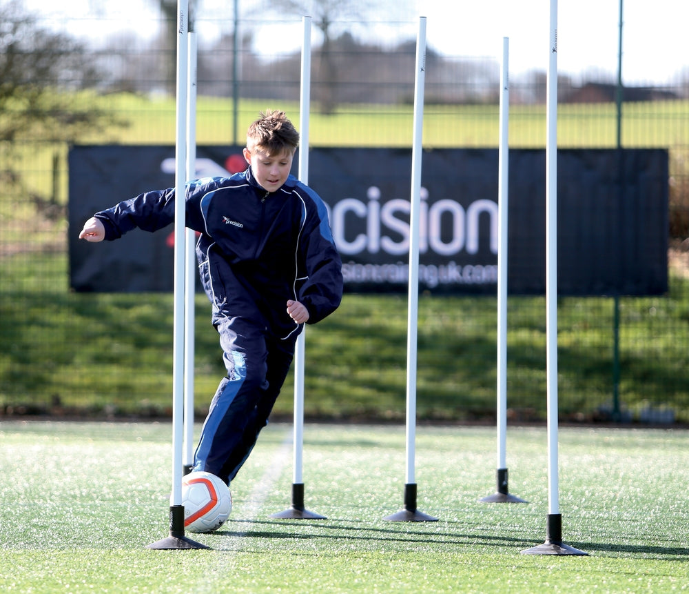 A boy in a navy tracksuit dribbles a soccer ball through Precision Training Boundary Poles (Set of 12), each secured with ground spikes, on a green outdoor field.