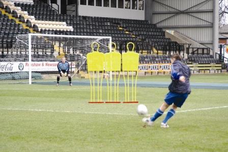 During practice, a soccer player aims the ball past a Precision Training Pro Mannequin from Precision Training as the goalkeeper prepares to defend in front of an empty stadium.