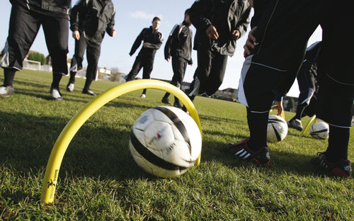 A group in black sportswear practices soccer drills on grass. A Precision Training Precision Spiked Passing Arc (Outdoor) aids their passing accuracy as a soccer ball rolls through it, with other players and balls visible in the background.