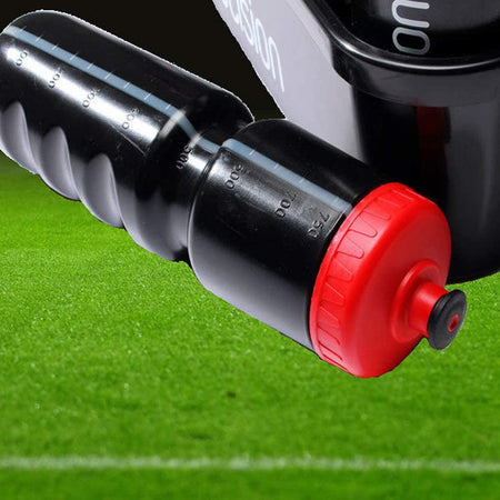 A close-up of the Precision Training Water Bottles & Carrier Black by Precision Training, 750ml with a red cap, lies on green grass near sports equipment and a white line marking is visible in the background.
