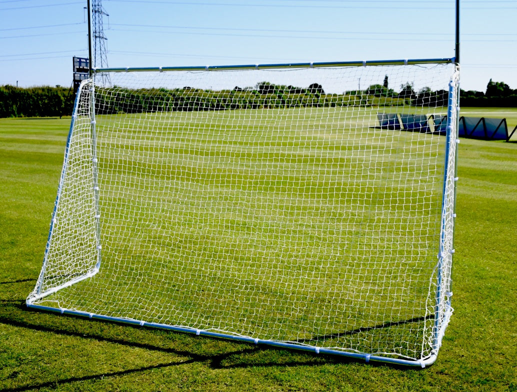 The Precision Training Precision Multi Sport Steel Goal with a zinc-coated frame is set up on a green grass field under a blue sky, with trees, a fence, and power lines visible in the background.