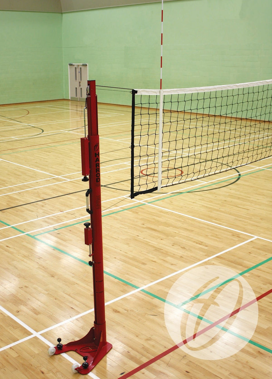 A Harrod Sport Antennae in Sheaths set is installed on a volleyball net in an empty indoor gym with wooden floors and colored court lines. The net is attached to a red pole.