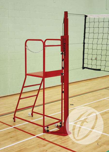 The Harrod Sport Steel Volleyball Referee Stand, featuring a non-slip platform and red finish, is placed next to a volleyball net on an indoor wooden court with light green walls and white boundary lines.