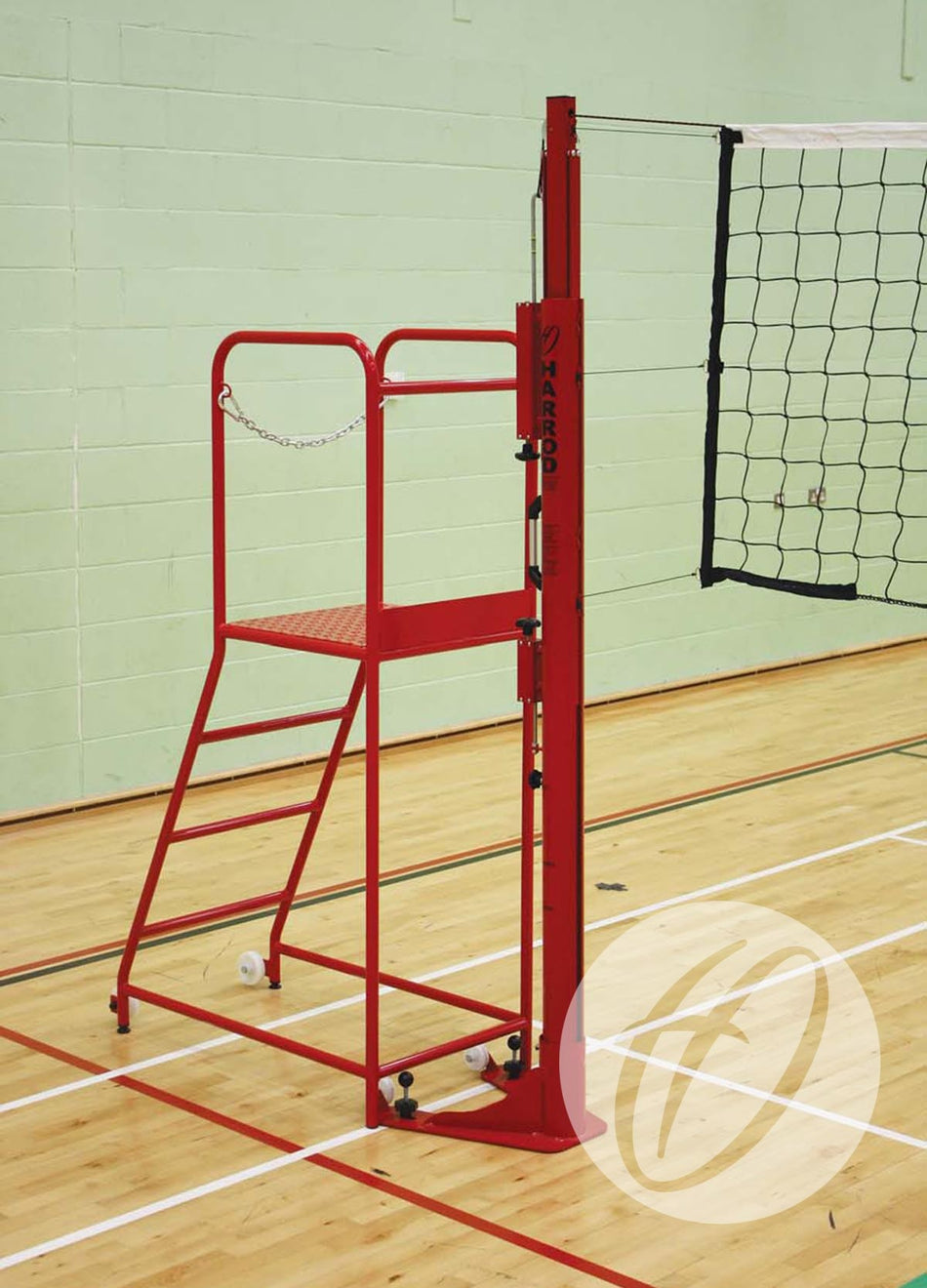 The Harrod Sport Steel Volleyball Referee Stand, featuring a non-slip platform and red finish, is placed next to a volleyball net on an indoor wooden court with light green walls and white boundary lines.
