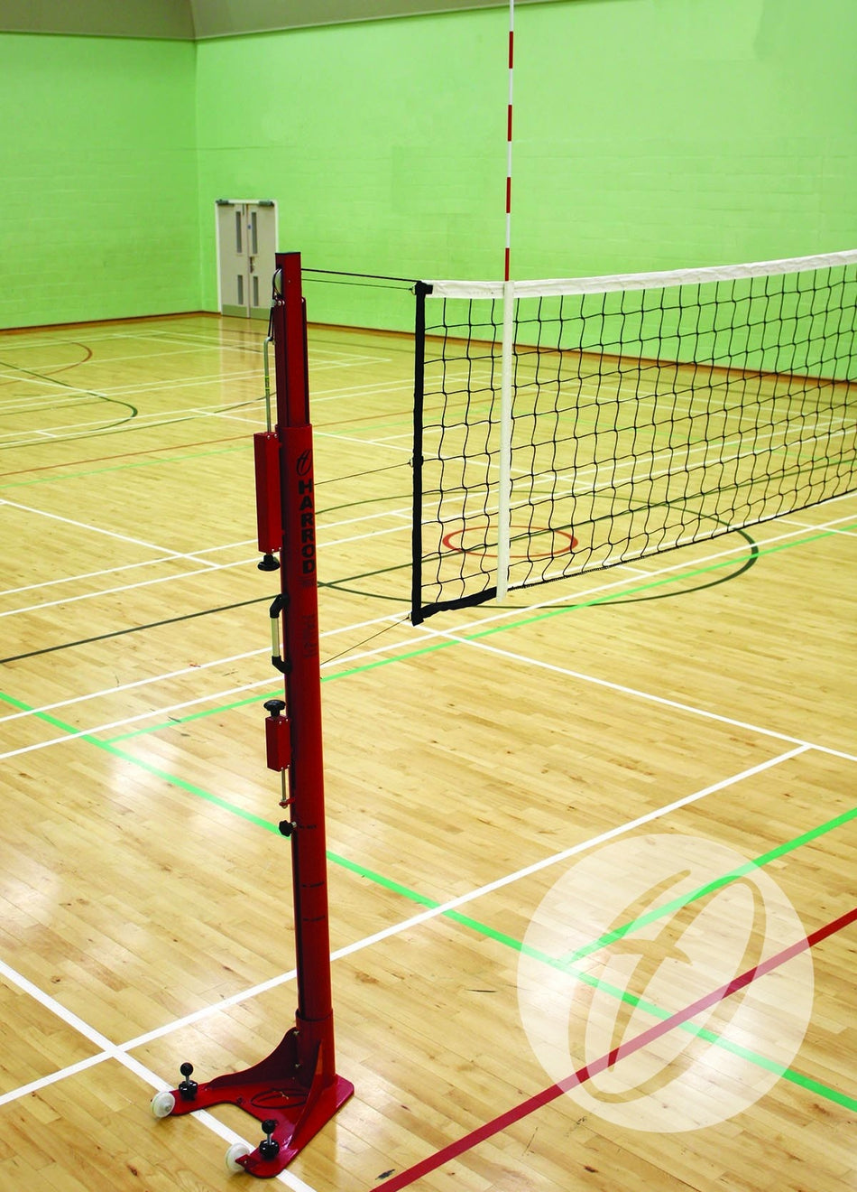 Harrod Sport Floor Fixed Competition Volleyball Posts are set up in a well-lit indoor gym with a wooden floor, green walls, and colorful court markings. The posts support an adjustable volleyball net for competitive play.