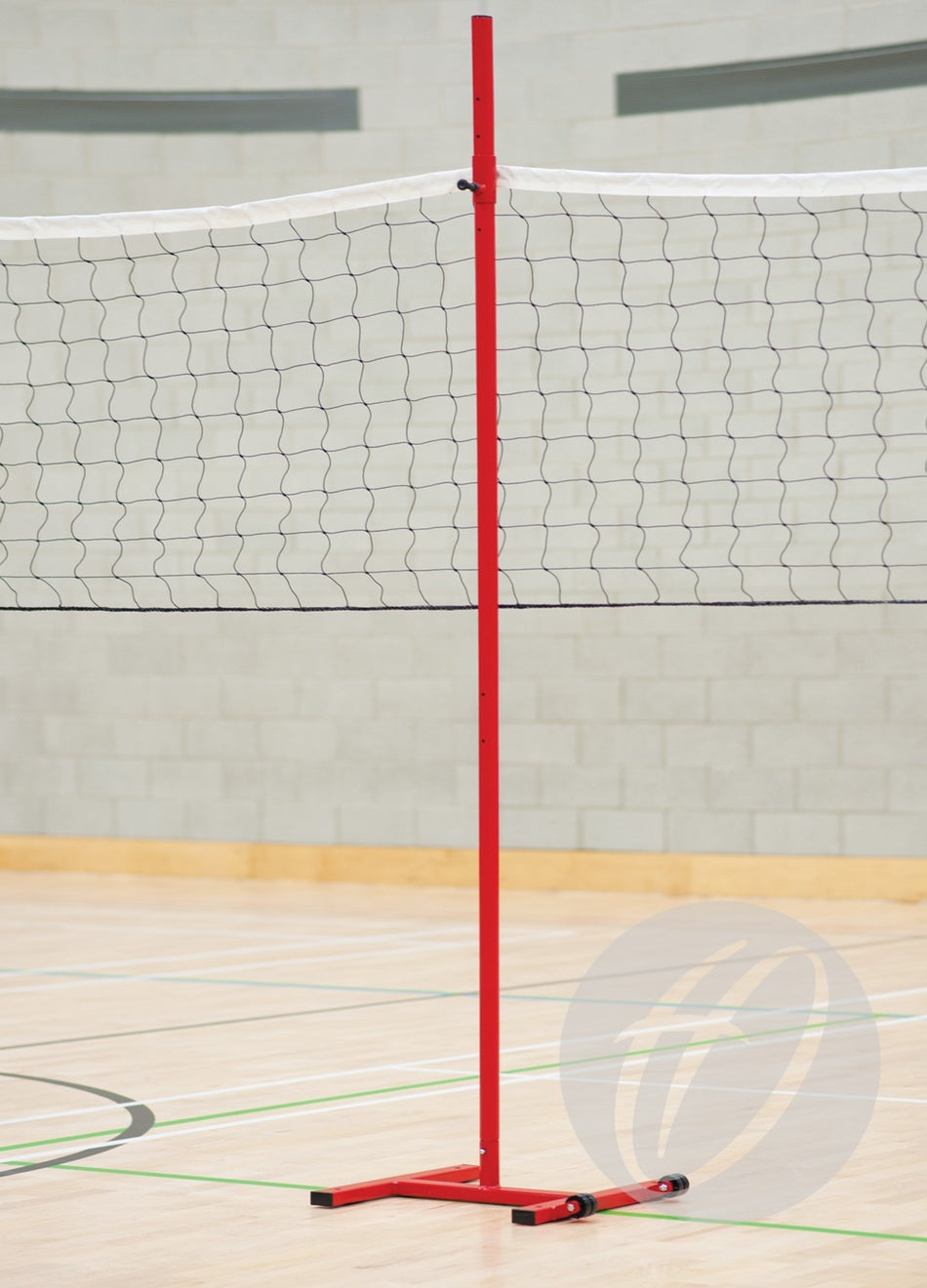 A Harrod Sport Intermediate Support Post holds a black and white volleyball net on a light wood indoor court with white and green floor markings, set against a pale brick wall backdrop.