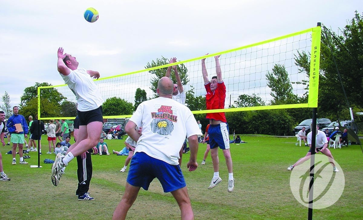 Players enjoy a game on grass with the Harrod Sport Portable Volleyball set. A player in white jumps to spike as two opponents in blue and red block at the net, while spectators watch nearby.