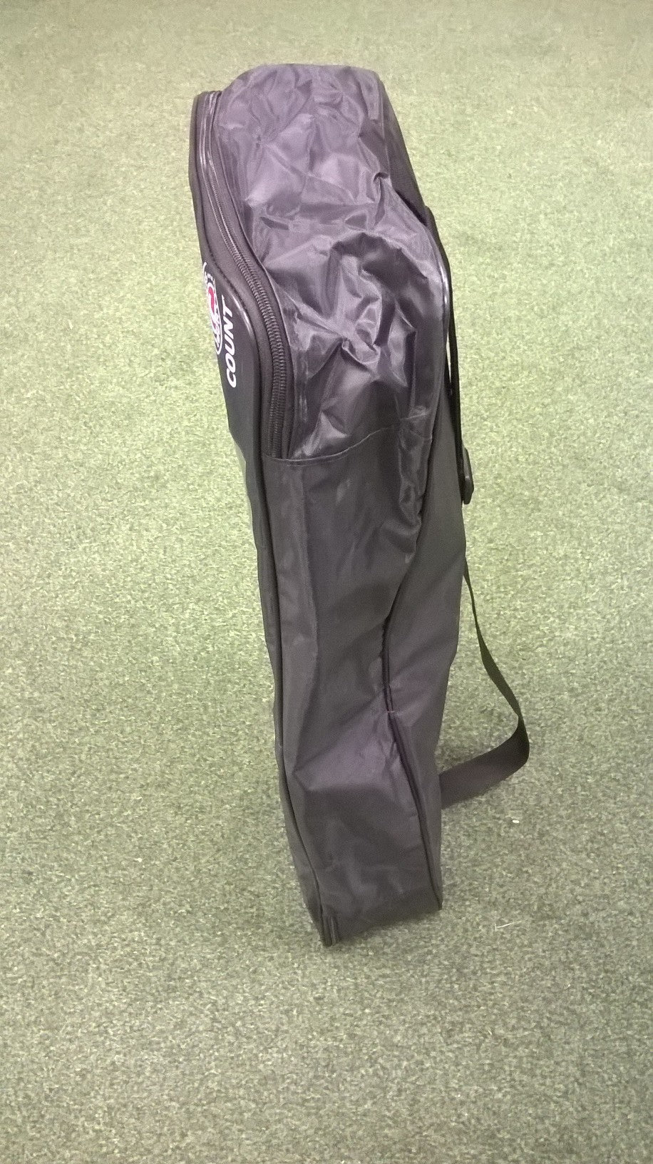 The Hunts County Beach Cricket Set comes in a tall, black sports bag with a zipper and shoulder strap, standing upright on green carpet—ideal for carrying your gear to the beach.