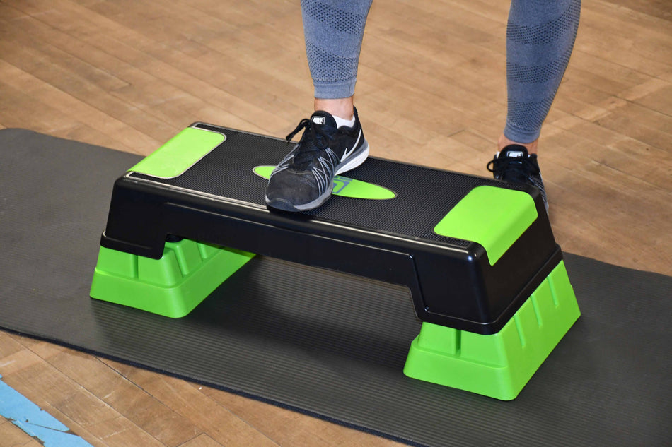 Wearing leggings and sneakers, a person steps onto the Urban Fitness Adjustable Aerobic Step by Urban Fitness, featuring a non-slip surface, set on a black exercise mat atop a wooden floor.