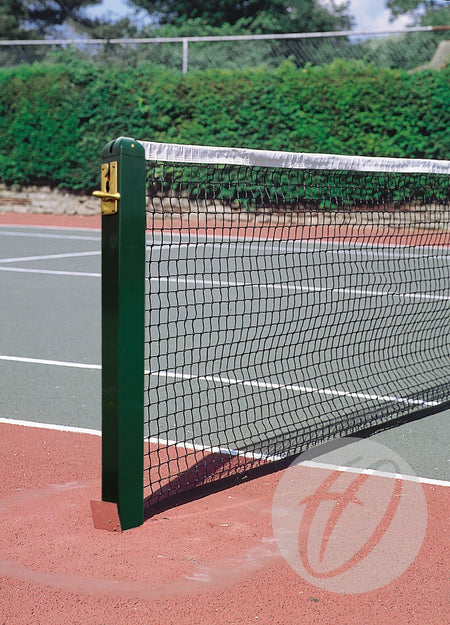 A close-up of Harrod Sport Aluminium Tennis Posts with locking sockets, installed on a red and green outdoor court; trees and a fence are visible in the background.