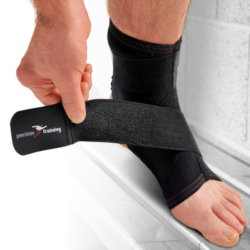 A person wraps the Precision Training Precision Neoprene Ankle with Strap Support, which is black and adjustable, around their right ankle for compression and support. Their foot rests on a light-colored surface.