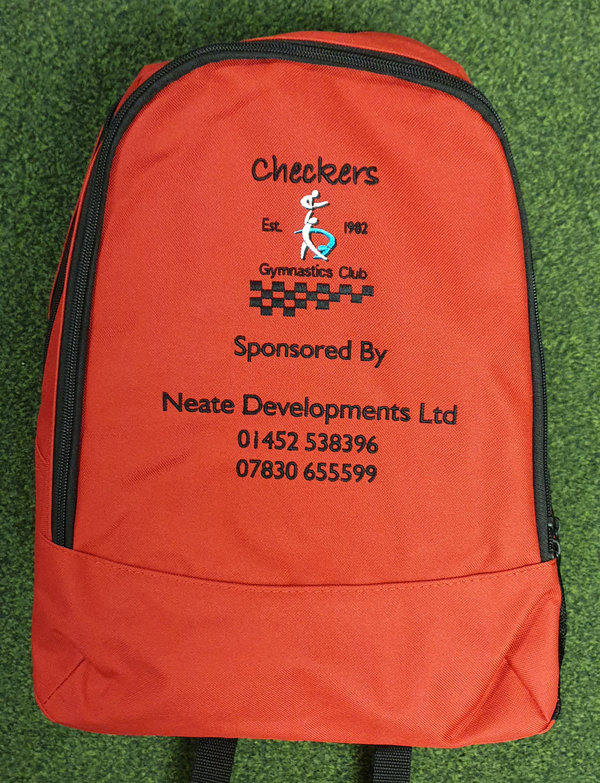 The PenCarrie Checkers Gymnastics Rucksack is a red 18L bag with embroidered club logo, sponsor details, and phone numbers in black and orange, plus a padded back panel, shown on green carpet.