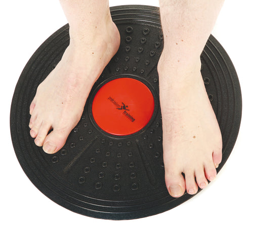 Two bare feet stand on the Precision Training Precision Balance Board, a round black board with a red center, made of heavy-duty plastic—ideal for balance training or sports rehabilitation.