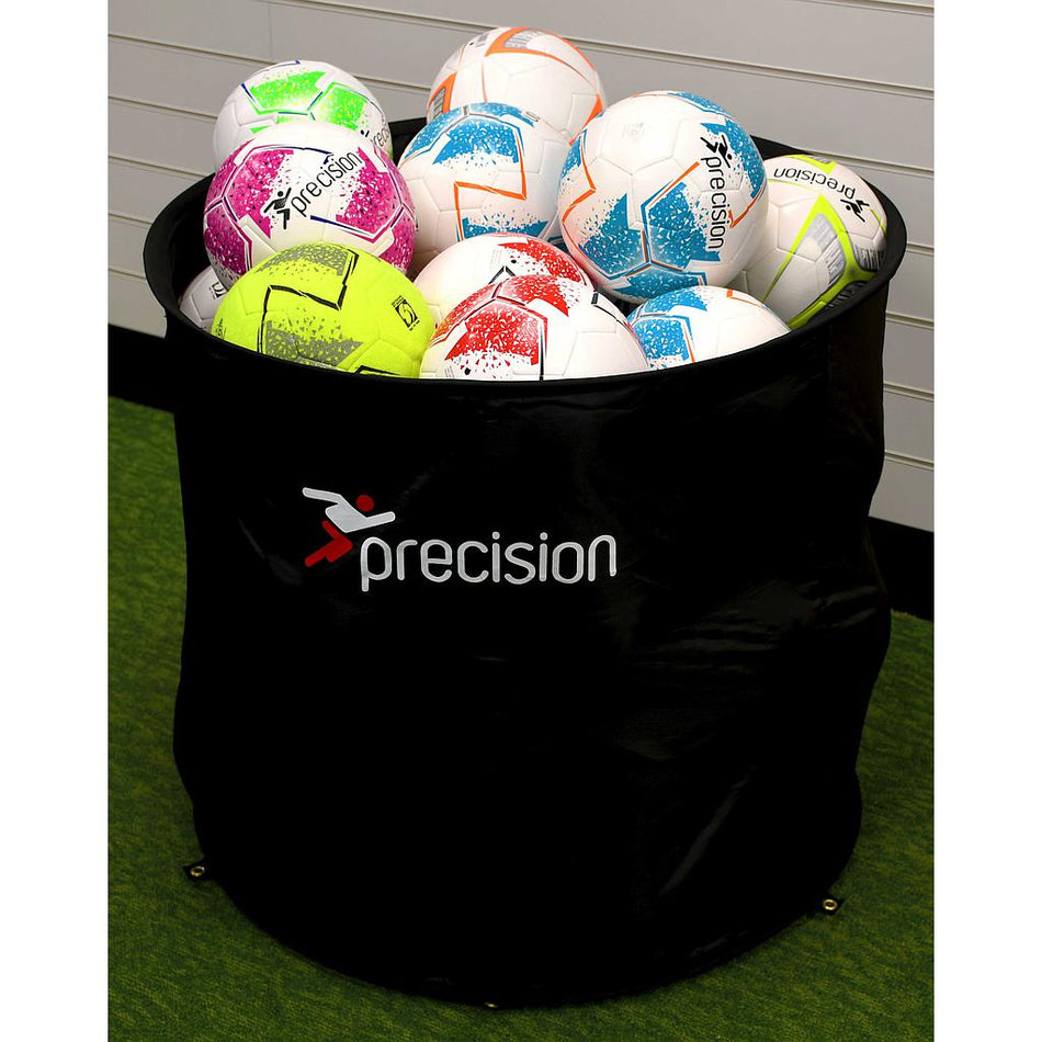 The Precision Training Ball Bin - POS/On Field, in black, is filled with vibrant soccer balls—white, pink, blue, green, orange, and yellow—and sits on a green floor against a wall with white paneling.