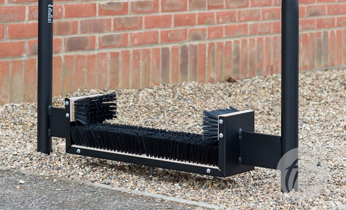 A Harrod Sport Socketed Premier Boot Wiper 1.1m, featuring side and bottom brushes, is installed on gravel beside a brick wall—making it ideal for communal changing areas or as a boot wiper to keep footwear clean.