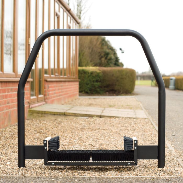 Installed on a gravel path beside a brick building with glass windows, the Harrod Sport Socketed Premier Boot Wiper 1.1m features sturdy metal construction and brushes on both sides. Perfect for communal changing areas, this practical fixture helps keep dirt at bay, with shrubs and trees visible in the background.