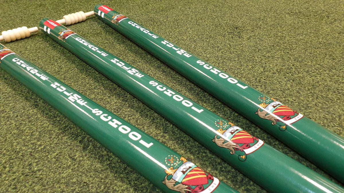 Three Gray Nicolls Custom Printed Club Cricket Stumps with colorful crests and "Foos On - Master Series" branding are arranged parallel on a textured green surface, evoking a personalised, premium cricket experience.