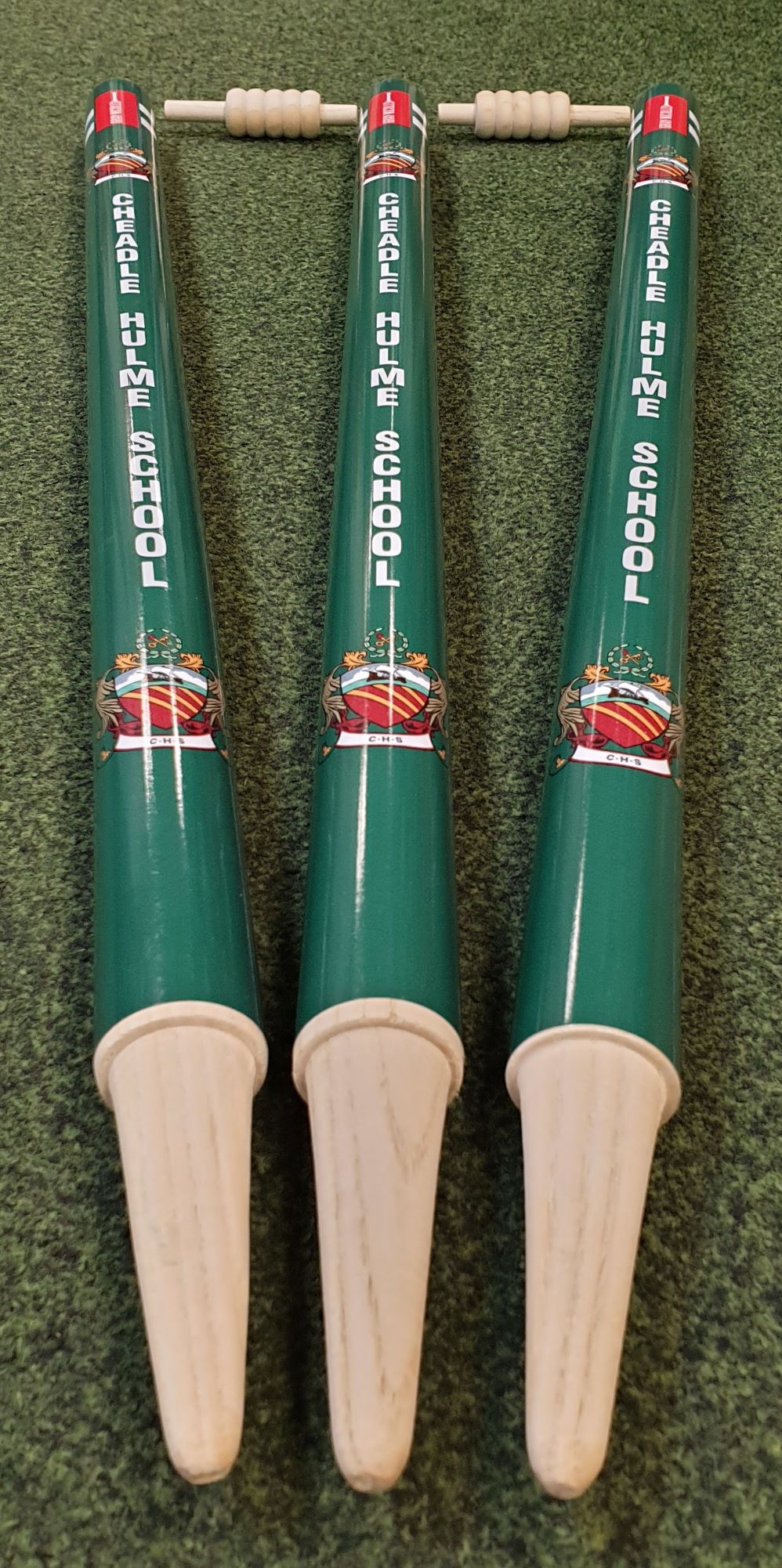 Three green and white Gray Nicolls Custom Printed Club Cricket Stumps, featuring Cheadle Hulme School’s name and crest, are displayed side by side on a green textured surface.