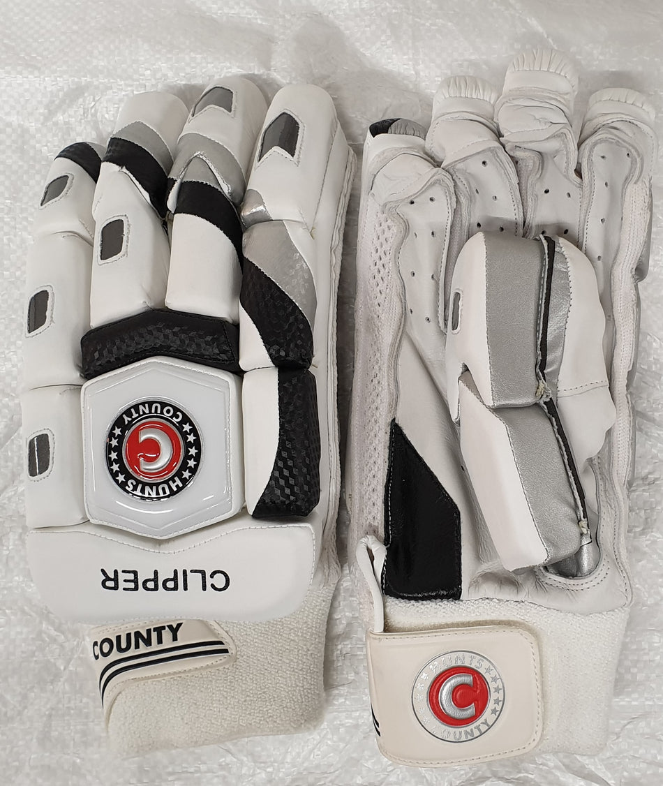 The Hunts County Clipper Batting Glove features a white split-finger design with black and silver accents, COUNTY branding, red and black wrist logo, calf leather palm, extra protection, and is shown on a textured white surface.