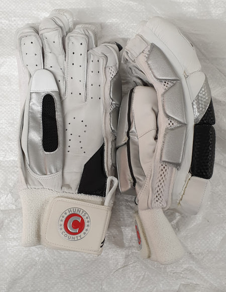 The Hunts County Clipper Batting Glove by Hunts County features a white and silver split finger design with black accents, a protective calf leather palm, and a wrist strap displaying the circular County Hunts logo with a red “C”.