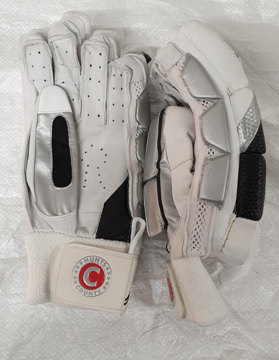 The Hunts County Clipper Batting Glove by Hunts County features a white and silver split finger design with black accents, a protective calf leather palm, and a wrist strap displaying the circular County Hunts logo with a red “C”.