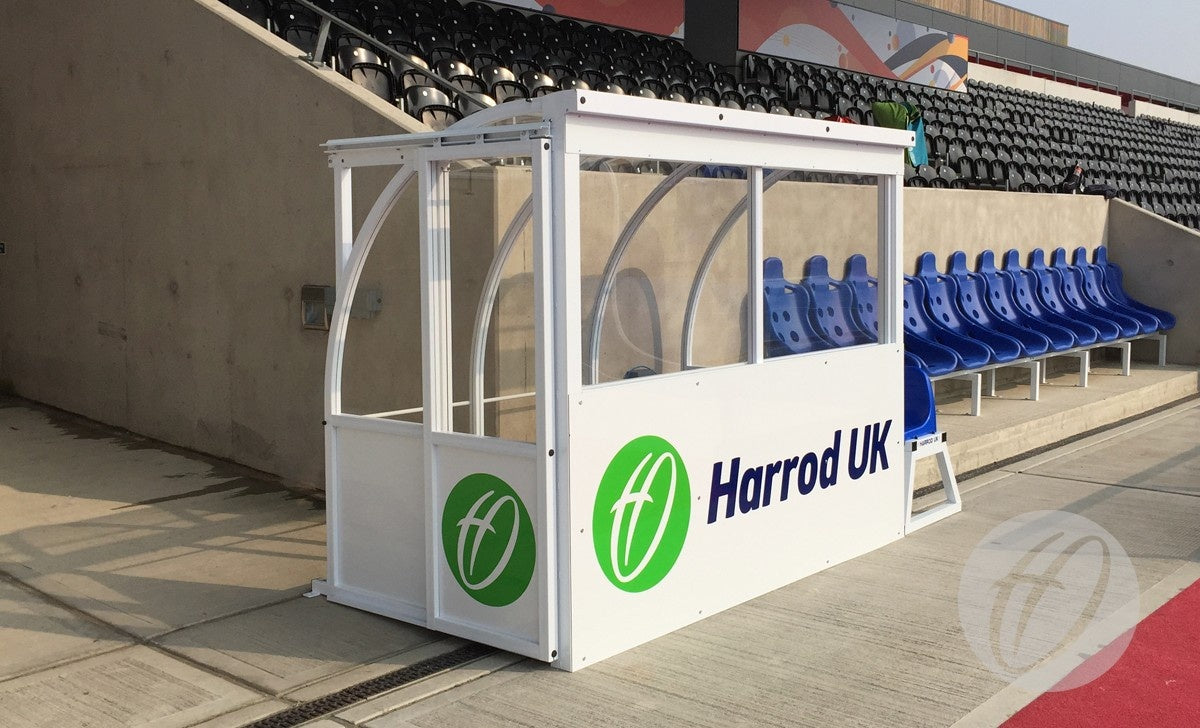 The Club Technical Shelter by Harrod Sport is a white and clear polycarbonate dugout with an aluminium panel and green logo, positioned next to blue stadium seats at an outdoor sports venue.