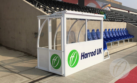 The Club Technical Shelter by Harrod Sport is a white and clear polycarbonate dugout with an aluminium panel and green logo, positioned next to blue stadium seats at an outdoor sports venue.