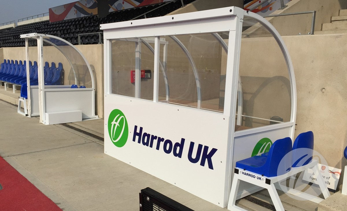 A Harrod Sport Club Technical Shelter with a clear polycarbonate viewing cover stands by blue stadium seats on concrete. Another similar shelter featuring an aluminium protective panel appears in the background.