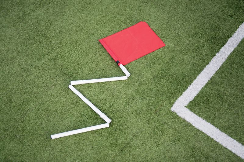 A Precision Training Precision Collapsible Corner Post from the set of 4 lies flat on green artificial turf by a white line, its bent zigzag pole showing how these usually sturdy posts can withstand tough impacts during play.
