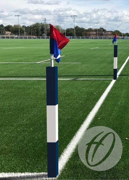 A rugby pitch with green artificial turf, white lines, and Harrod Sport 30MM Corner Pole Protection Pads on each corner post—topped with red and blue flags. The posts feature PVC high-density foam. Buildings and trees stand under a partly cloudy sky.