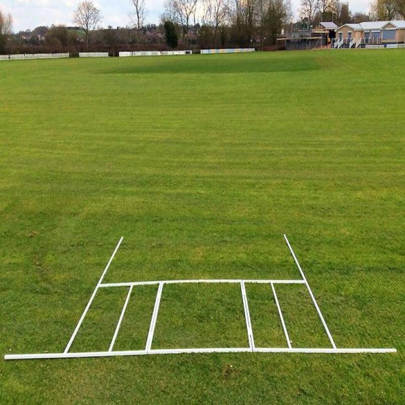 A Martin Berrill Sports Crease Marker with a folding steel frame outlines the cricket pitch on grass, with trees, a pavilion, and buildings in the background beneath a partly cloudy sky.