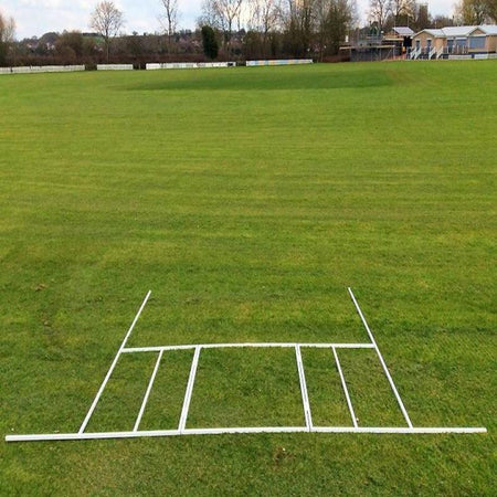 A Martin Berrill Sports Crease Marker with a folding steel frame outlines the cricket pitch on grass, with trees, a pavilion, and buildings in the background beneath a partly cloudy sky.