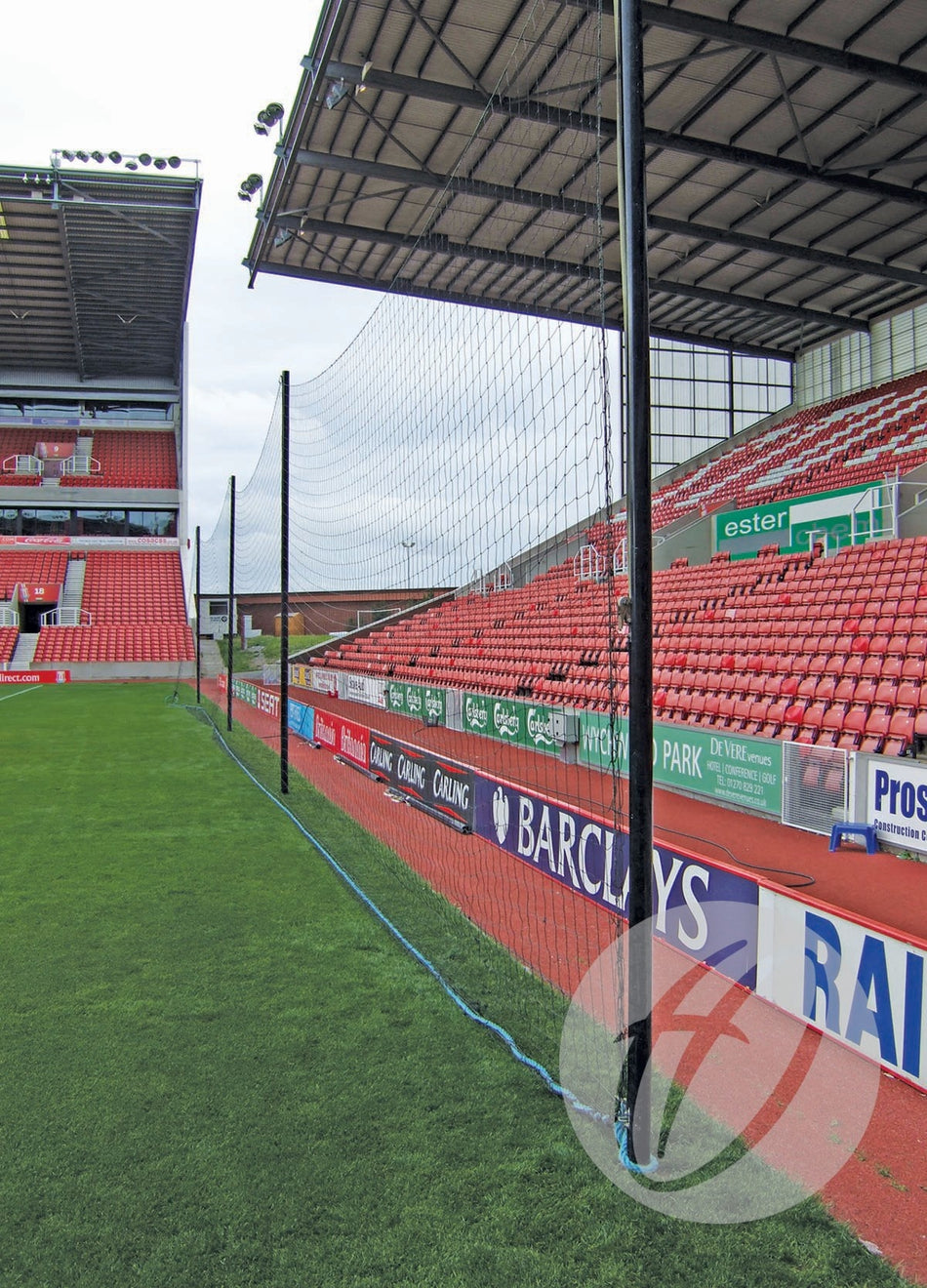 The Harrod Sport Crowd Protection Ball Stop System (80m x 5m), supported by strong aluminium poles, is installed along the soccer field, separating the green grass from empty red stadium seats and advertising boards.