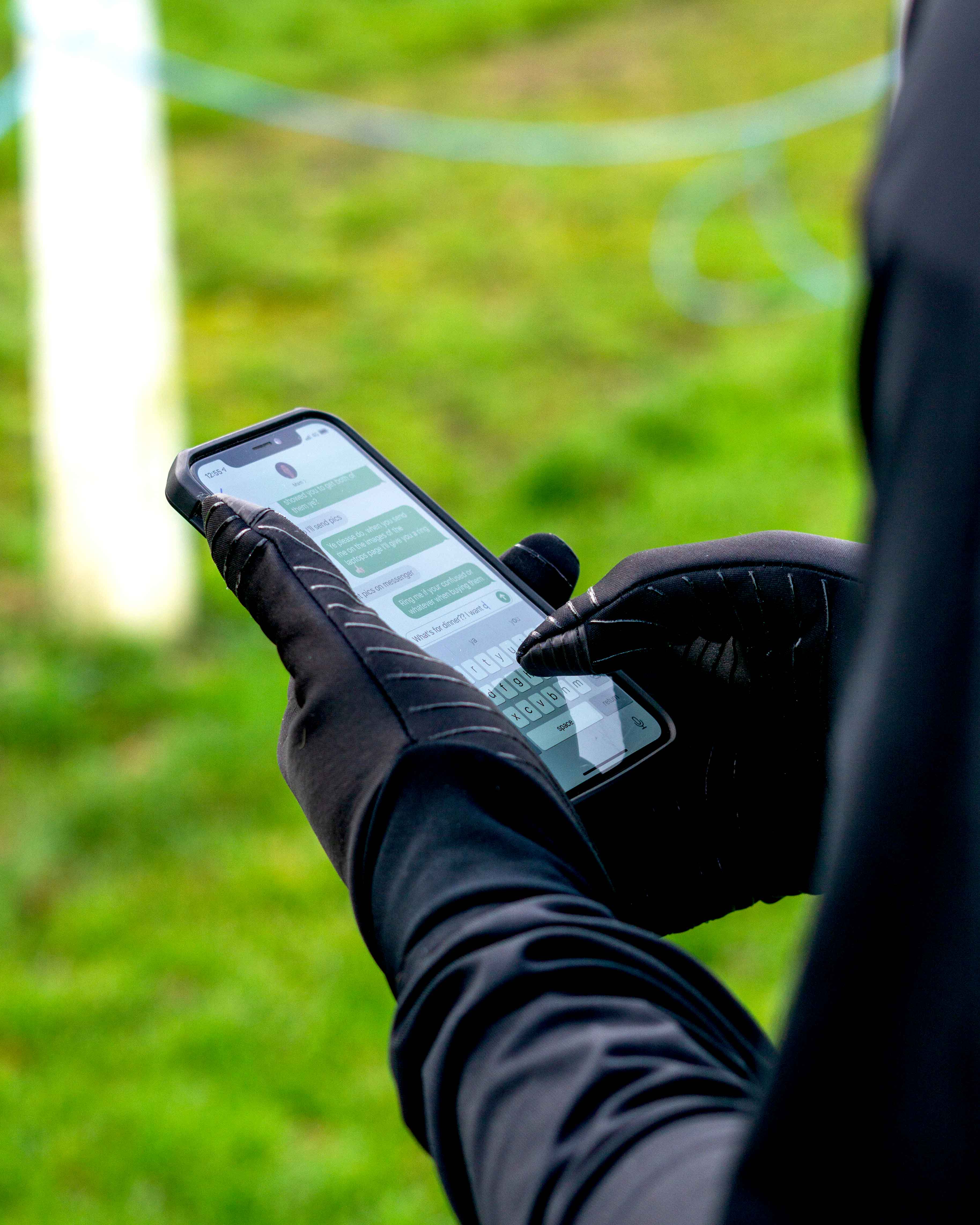 A person wearing Precision Essential Warm Players Gloves Adult by Precision Training and a dark jacket is texting on a smartphone, with a grassy field in the background.