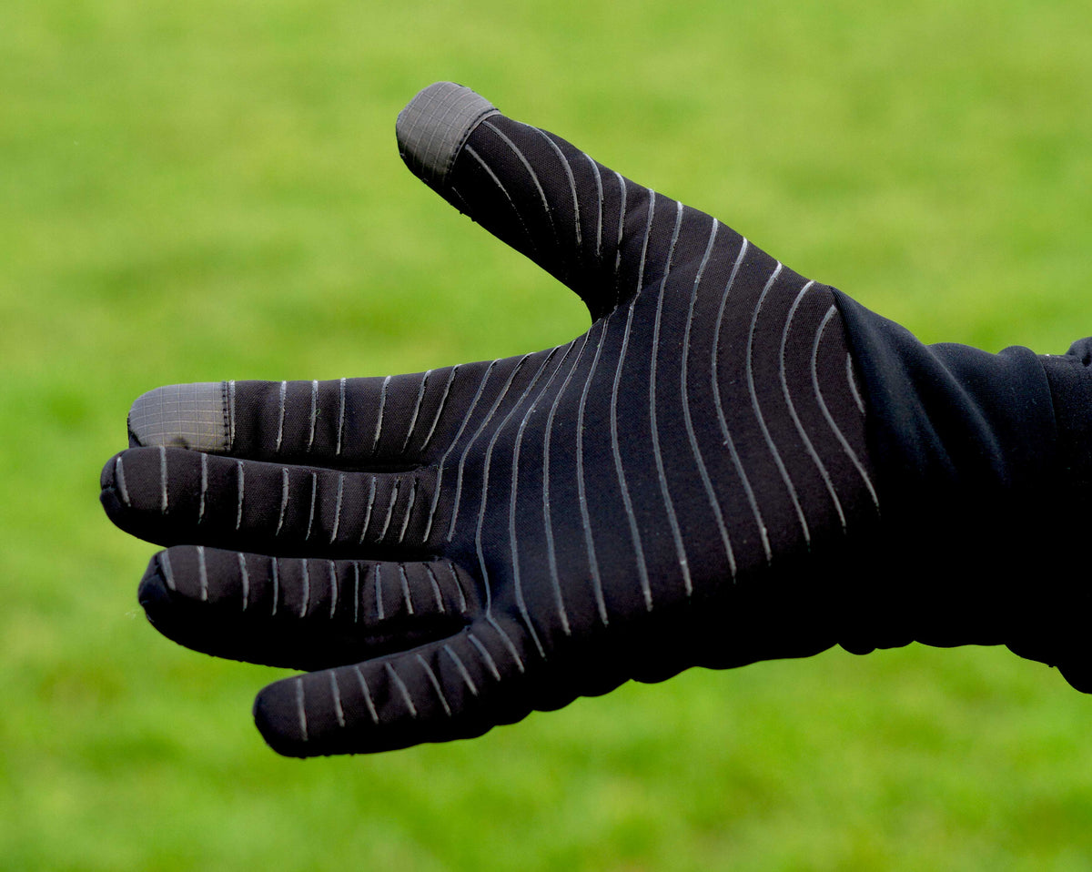A hand wears Precision Training Precision Essential Warm Players Gloves Adult with black fabric, thin white stripes, gray-tipped index finger and thumb, and a silicone palm grip, extended before a blurred green background.