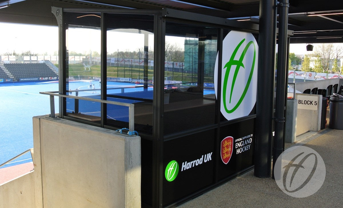 A Harrod Sport European Technical Shelter overlooks a blue hockey field in the stadium, displaying Harrod Sport and England Hockey logos, with empty spectator seats nearby.