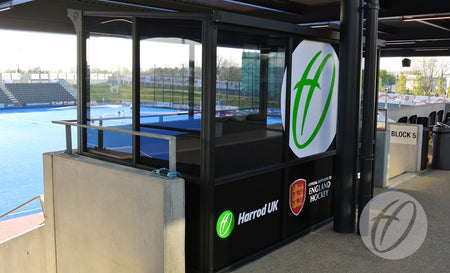 A Harrod Sport European Technical Shelter overlooks a blue hockey field in the stadium, displaying Harrod Sport and England Hockey logos, with empty spectator seats nearby.
