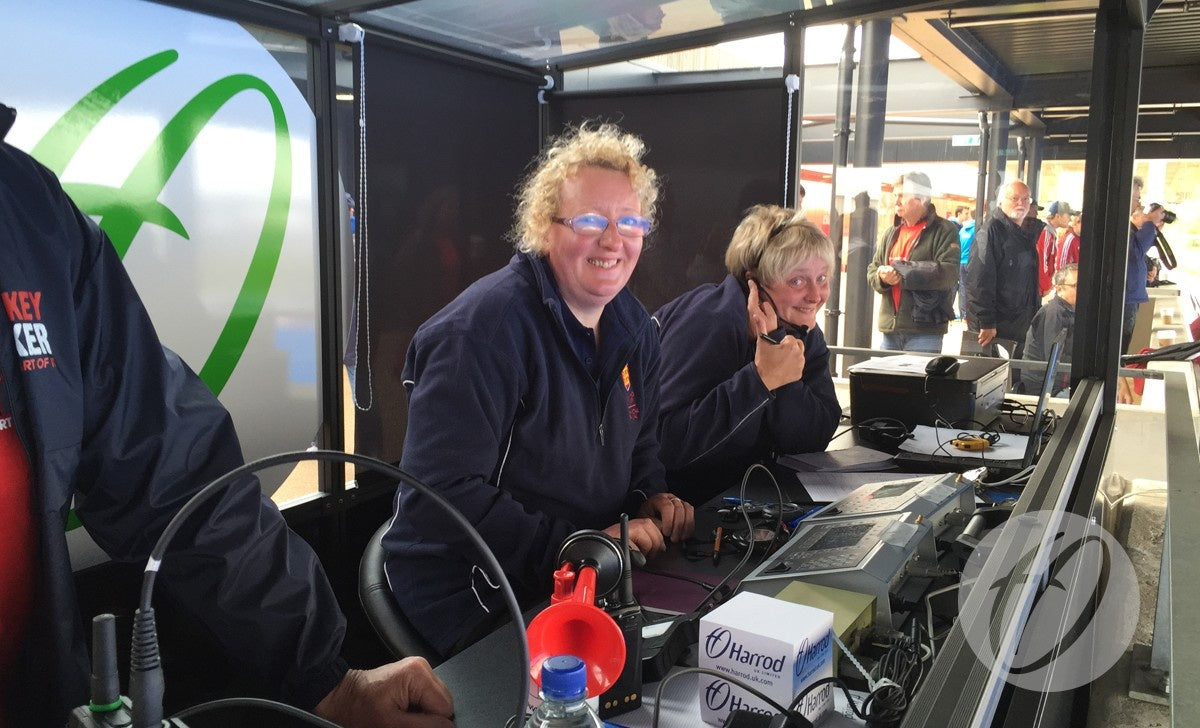 Two women sit at a control station in a Harrod Sport European Technical Shelter, smiling and giving thumbs-up. Wearing headsets and surrounded by equipment, they enjoy the lively atmosphere with others visible in the background.
