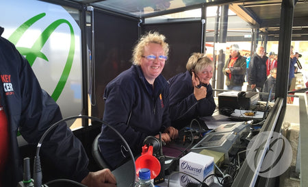 Two women sit at a control station in a Harrod Sport European Technical Shelter, smiling and giving thumbs-up. Wearing headsets and surrounded by equipment, they enjoy the lively atmosphere with others visible in the background.