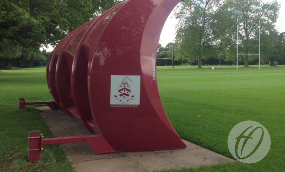 A curved, maroon Harrod Sport Fibretech "Pro" Shelter Anchor with the Bromsgrove School logo stands beside a grassy sports field with rugby goalposts and sturdy supports, surrounded by trees.
